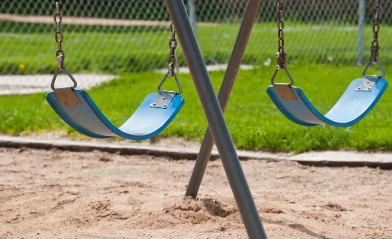 Two empty blue swings hanging on a playground swing set over sand with green grass in the background