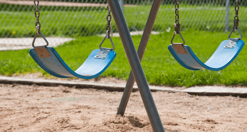 Two empty blue swings hanging on a playground swing set over sand with green grass in the background